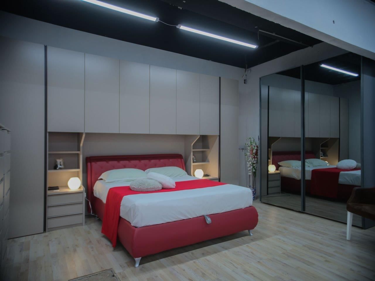 Una camera da letto moderna con letto rosso, armadi a muro color crema, illuminazione calda e un grande specchio che riflette il letto.