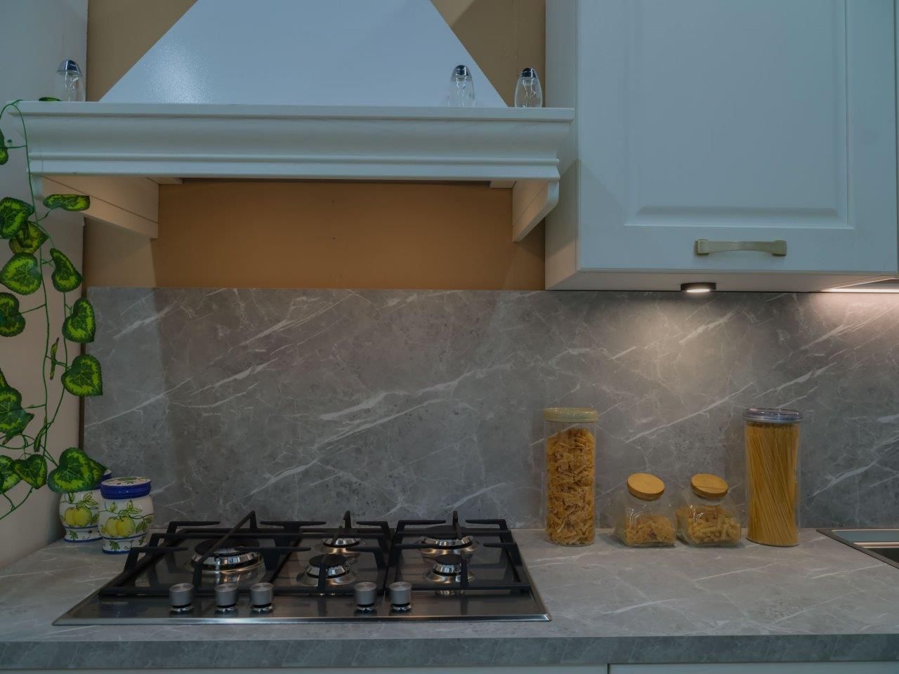 Una cucina moderna con piano cottura a gas, paraschizzi effetto marmo e barattoli di vetro su un piano di lavoro grigio.