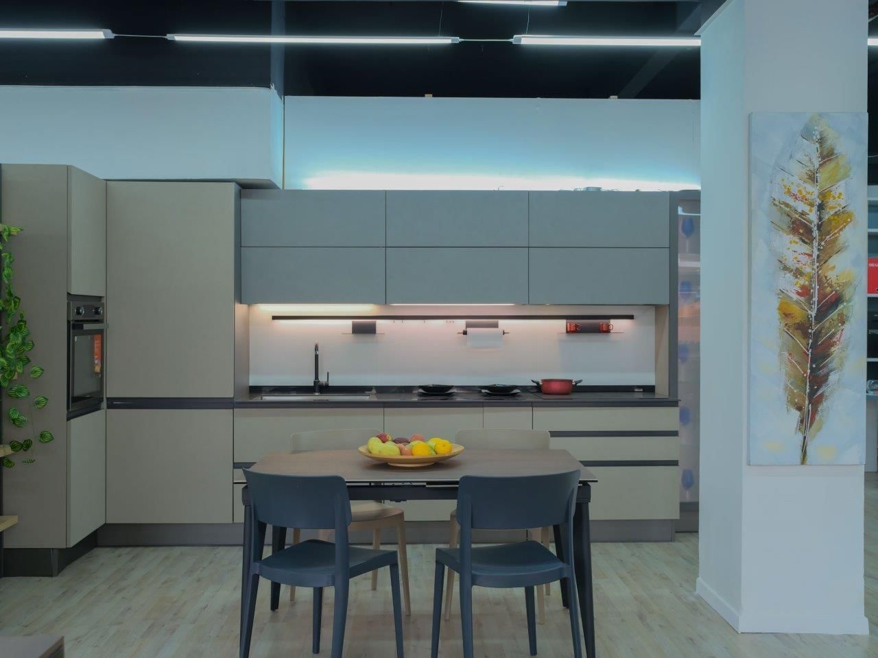 Cucina moderna con mobili beige, piano di lavoro scuro, tavolo da pranzo con due sedie e un quadro a forma di foglia.