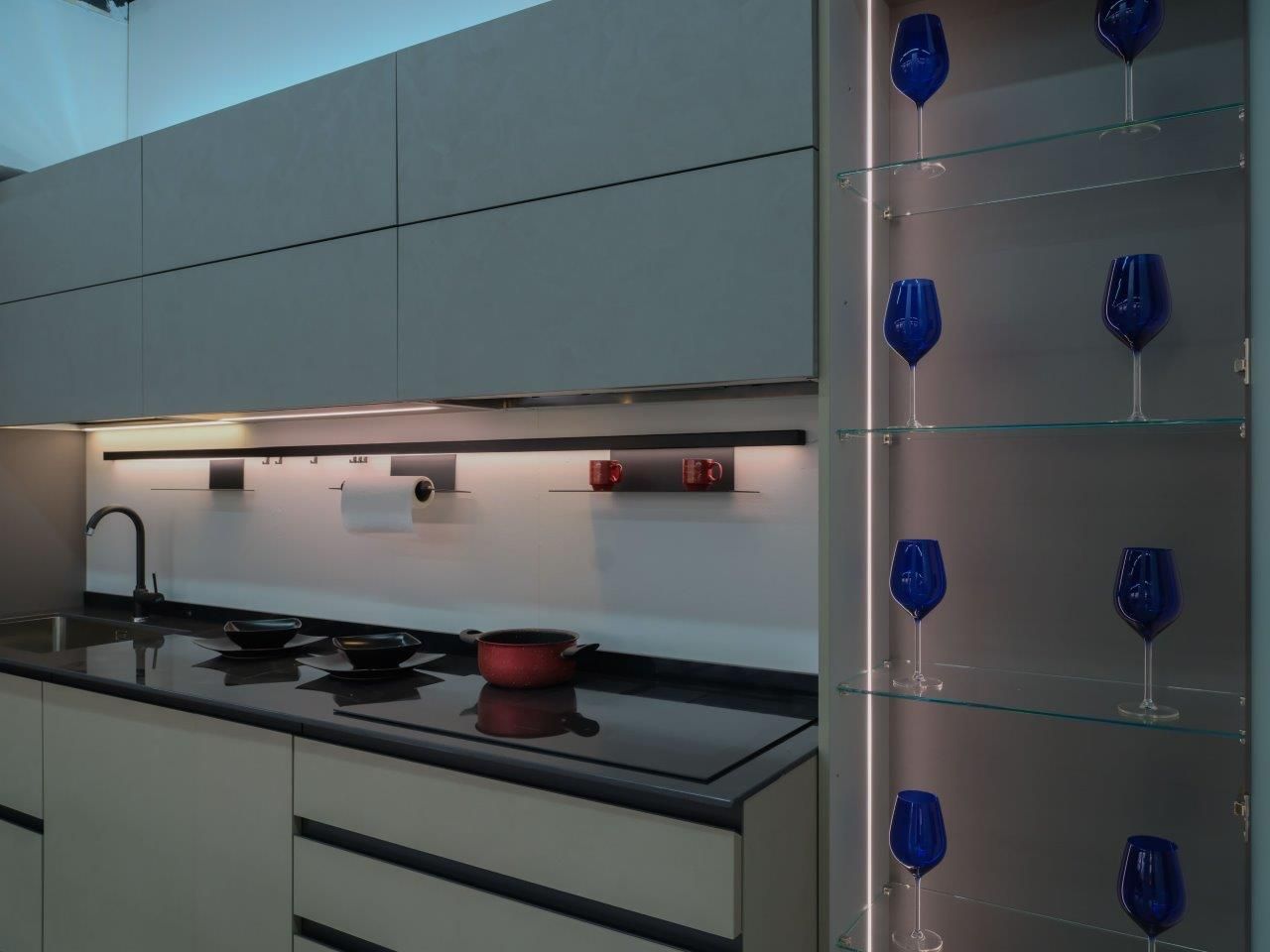 Cucina moderna con mobili grigi, un elegante piano di lavoro nero e una vetrina illuminata con calici da vino blu.