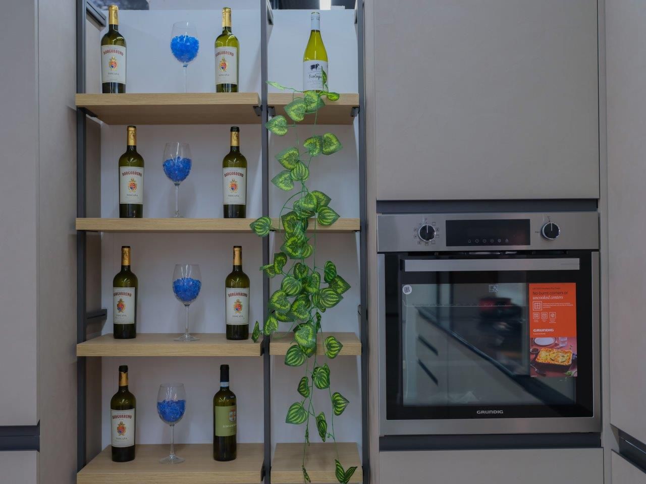 Scaffale con bottiglie di vino e bicchieri blu accanto a un forno a incasso, con una pianta di edera verde rampicante.