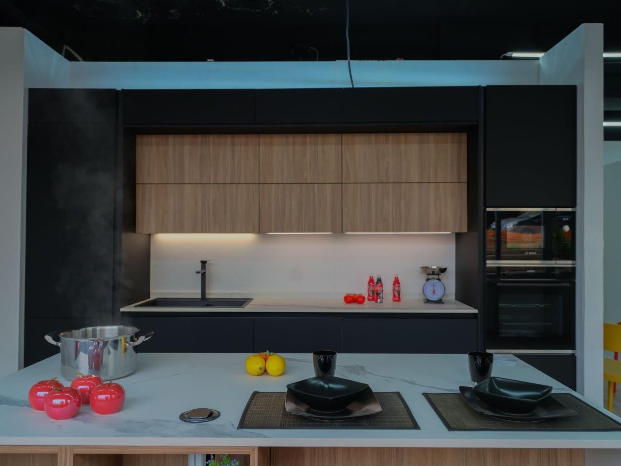Una cucina moderna con mobili in legno, dettagli neri, un'isola bianca, una pentola fumante, pomodori rossi e la tavola apparecchiata.
