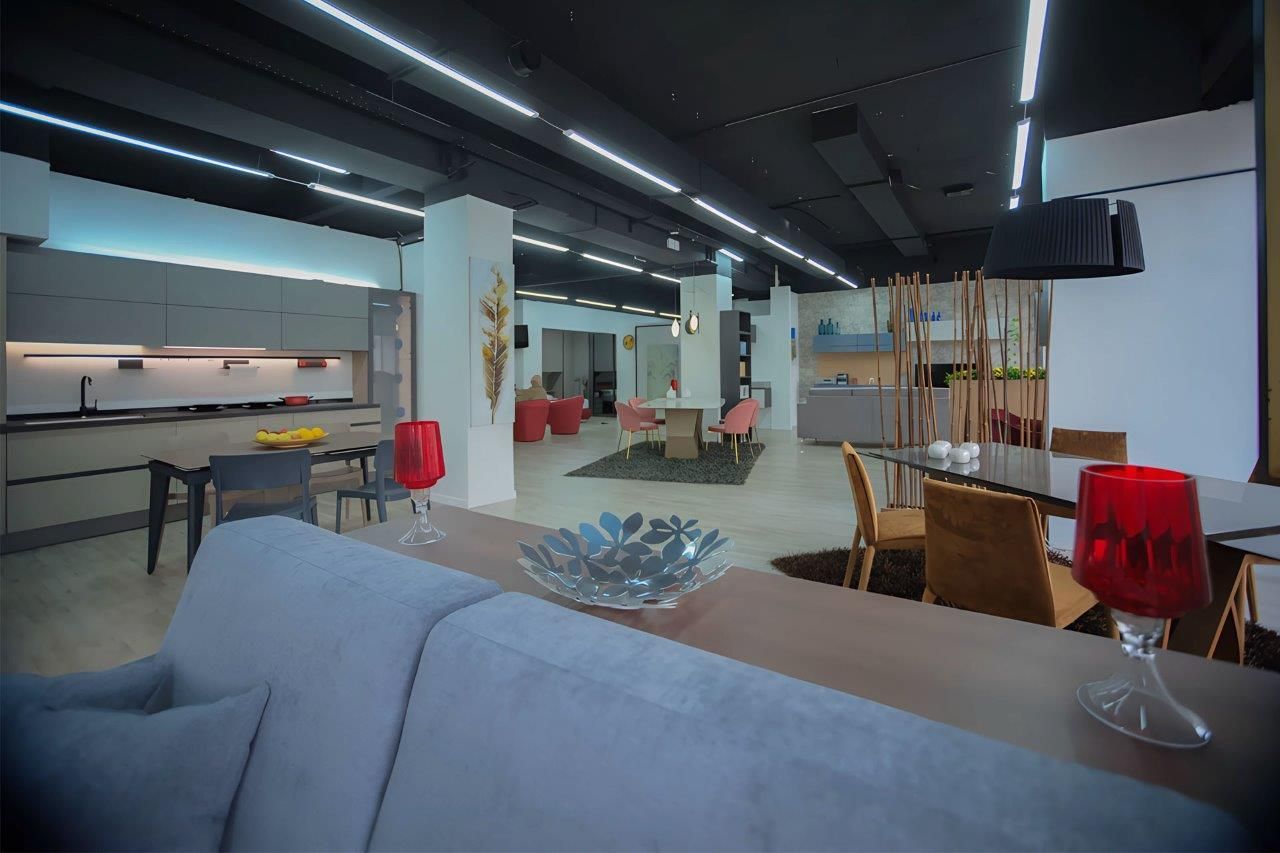 Interno di uno showroom di mobili moderni con cucine in esposizione, set da pranzo e un divano blu in primo piano.