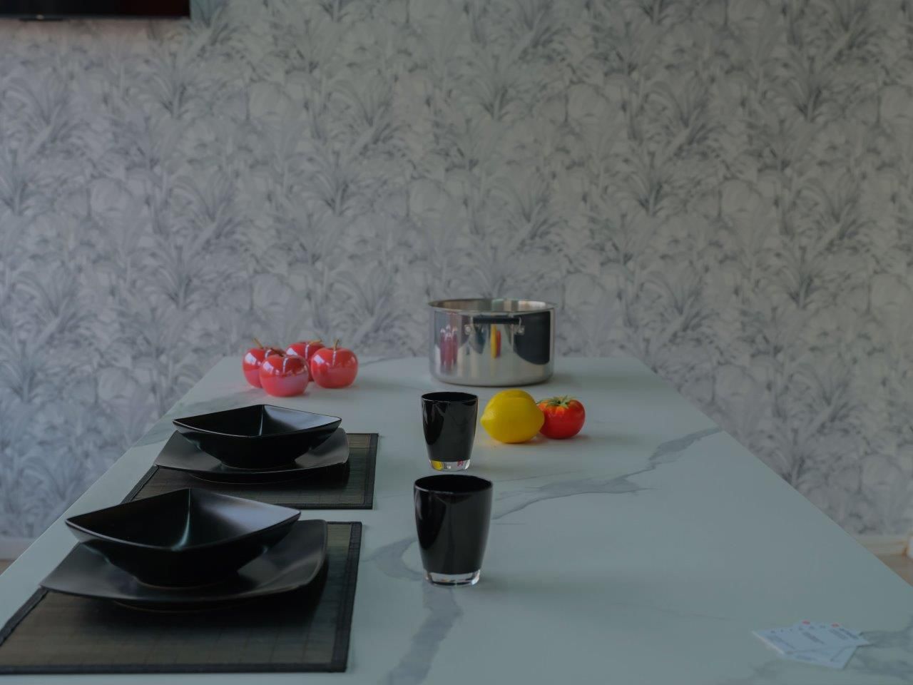 Un tavolo da pranzo con due posti a sedere, ciotole nere, bicchieri, una pentola di metallo e frutta, su uno sfondo di carta da parati a motivi geometrici.
