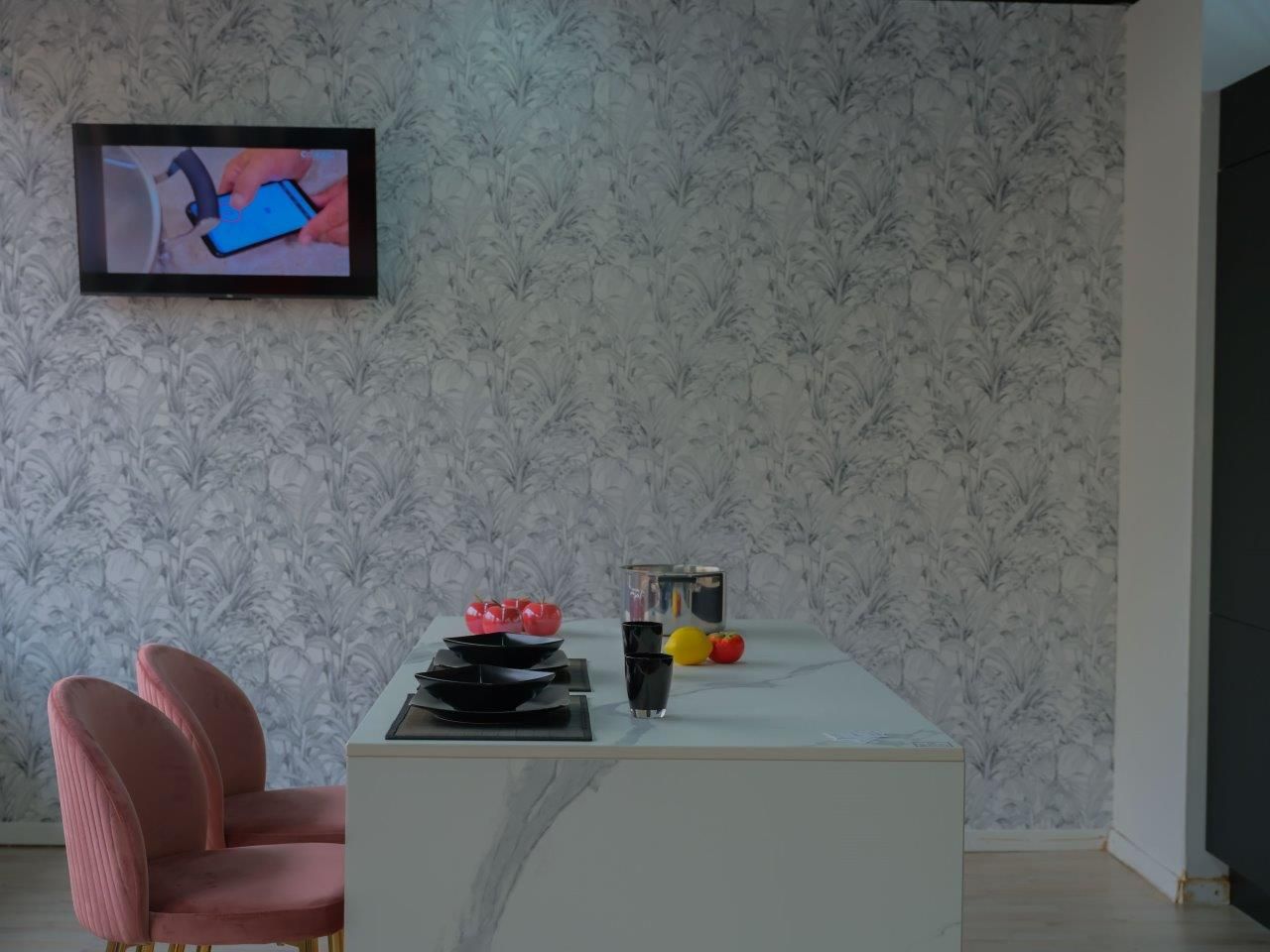 Una zona pranzo con un tavolo bianco e sedie rosa, su uno sfondo di carta da parati, con un televisore montato a parete.
