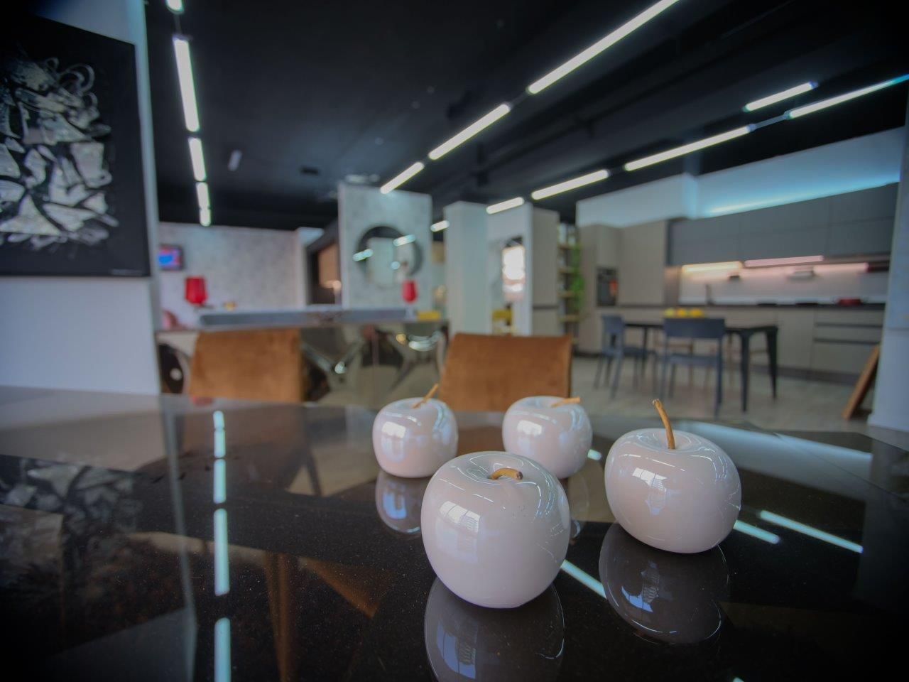 All'interno di uno showroom di cucine moderne a pianta aperta, tre mele decorative in ceramica bianca sono posizionate su un tavolo da pranzo scuro e riflettente.