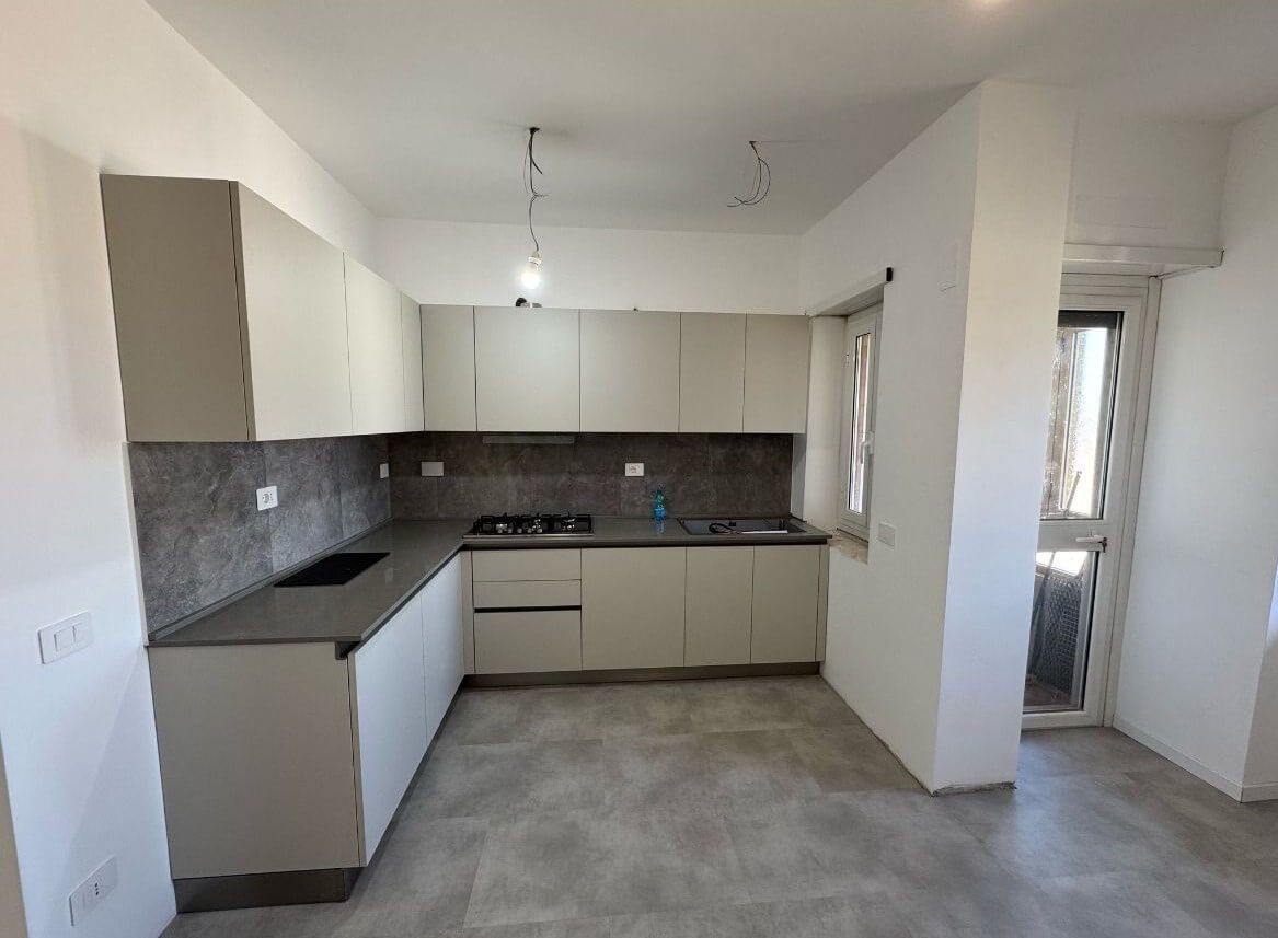 Cucina moderna a forma di L con mobili dai toni neutri, ripiani e pavimento piastrellati in grigio e una porta a vetri adiacente.