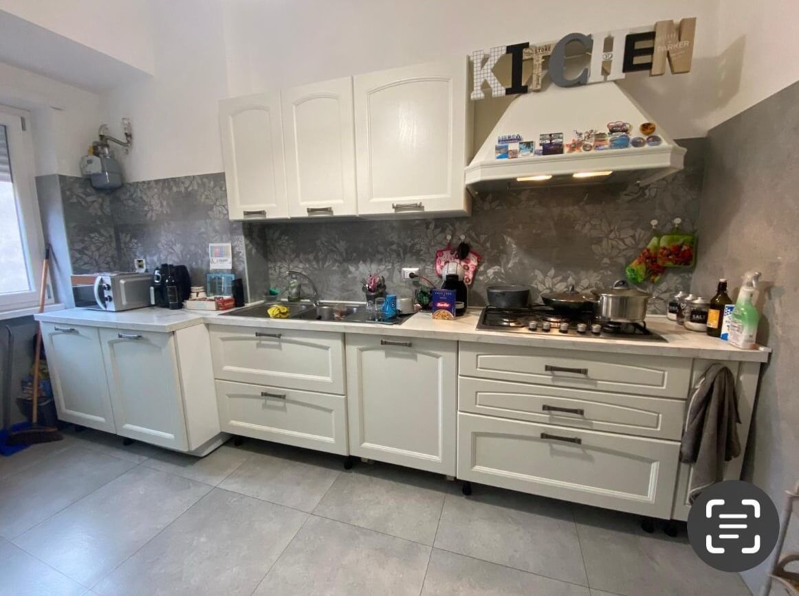 Una cucina dai colori chiari con mobili bianchi, un paraschizzi piastrellato, un piano cottura con cappa aspirante e un lavello.