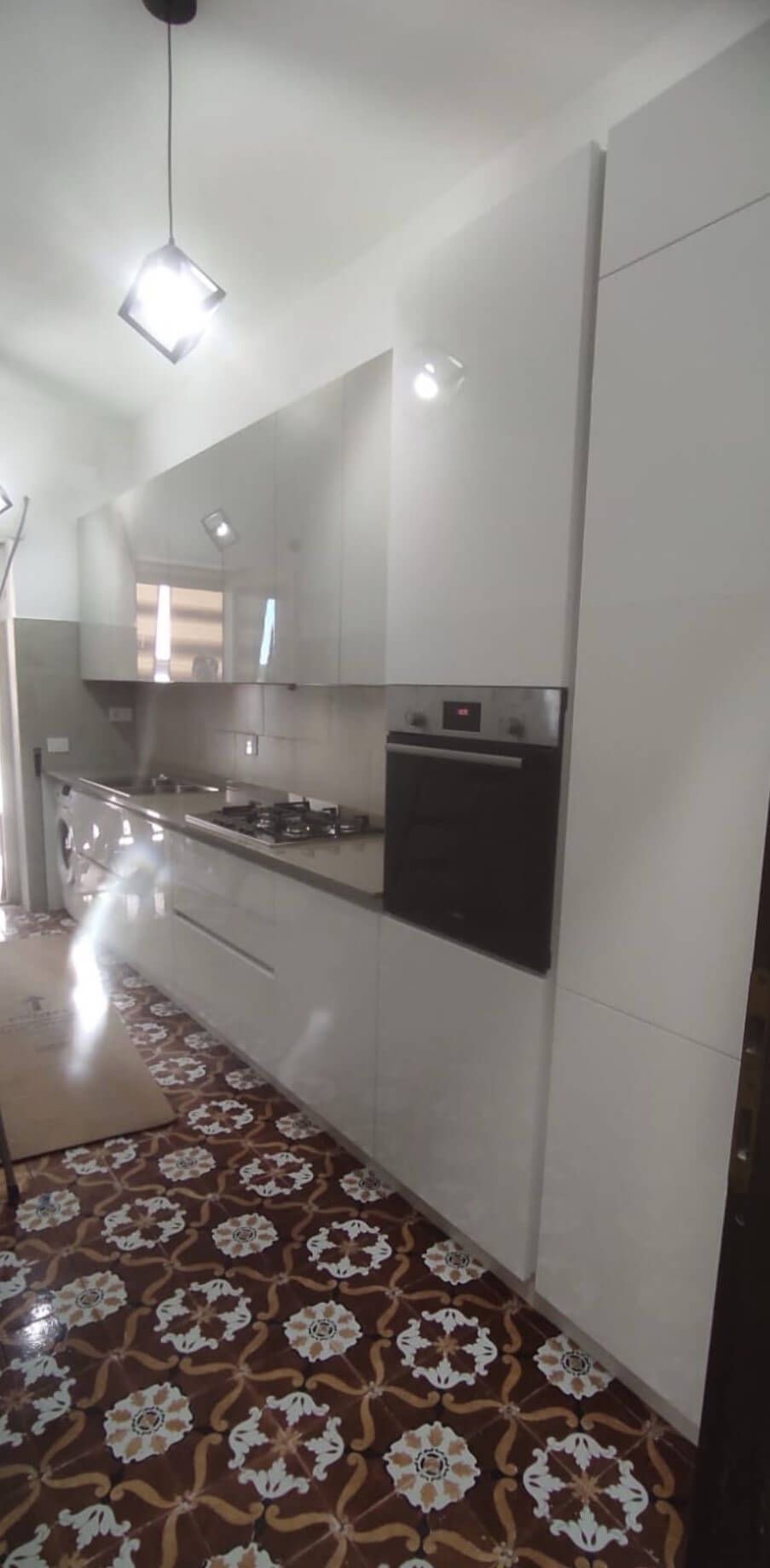 Una cucina moderna con mobili bianchi lucidi, forno a incasso e pavimento in piastrelle decorate e multicolori.