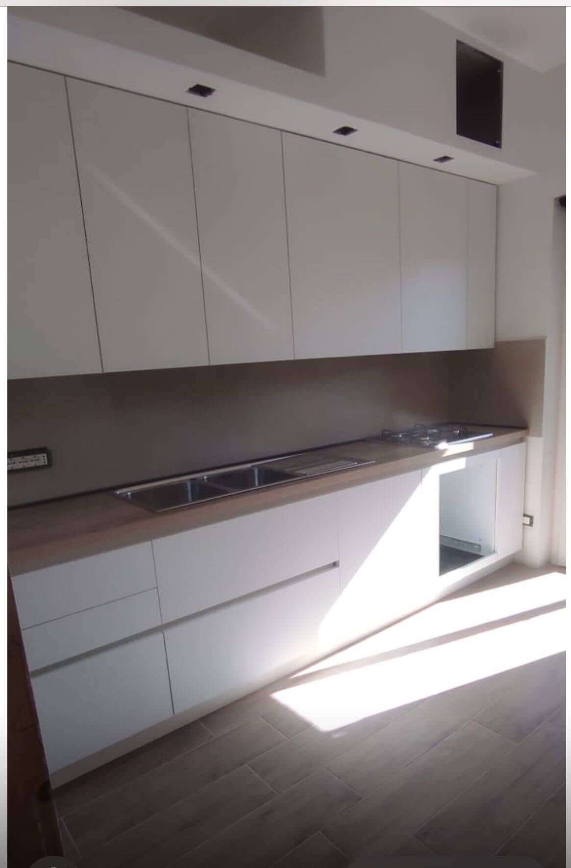 Una cucina moderna con mobili bianchi, piano di lavoro in legno chiaro e paraschizzi color tortora, in una stanza luminosa.