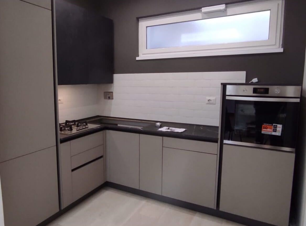 Cucina moderna a forma di L con mobili grigio chiaro, ripiani neri, forno in acciaio inox e paraschizzi piastrellato bianco.