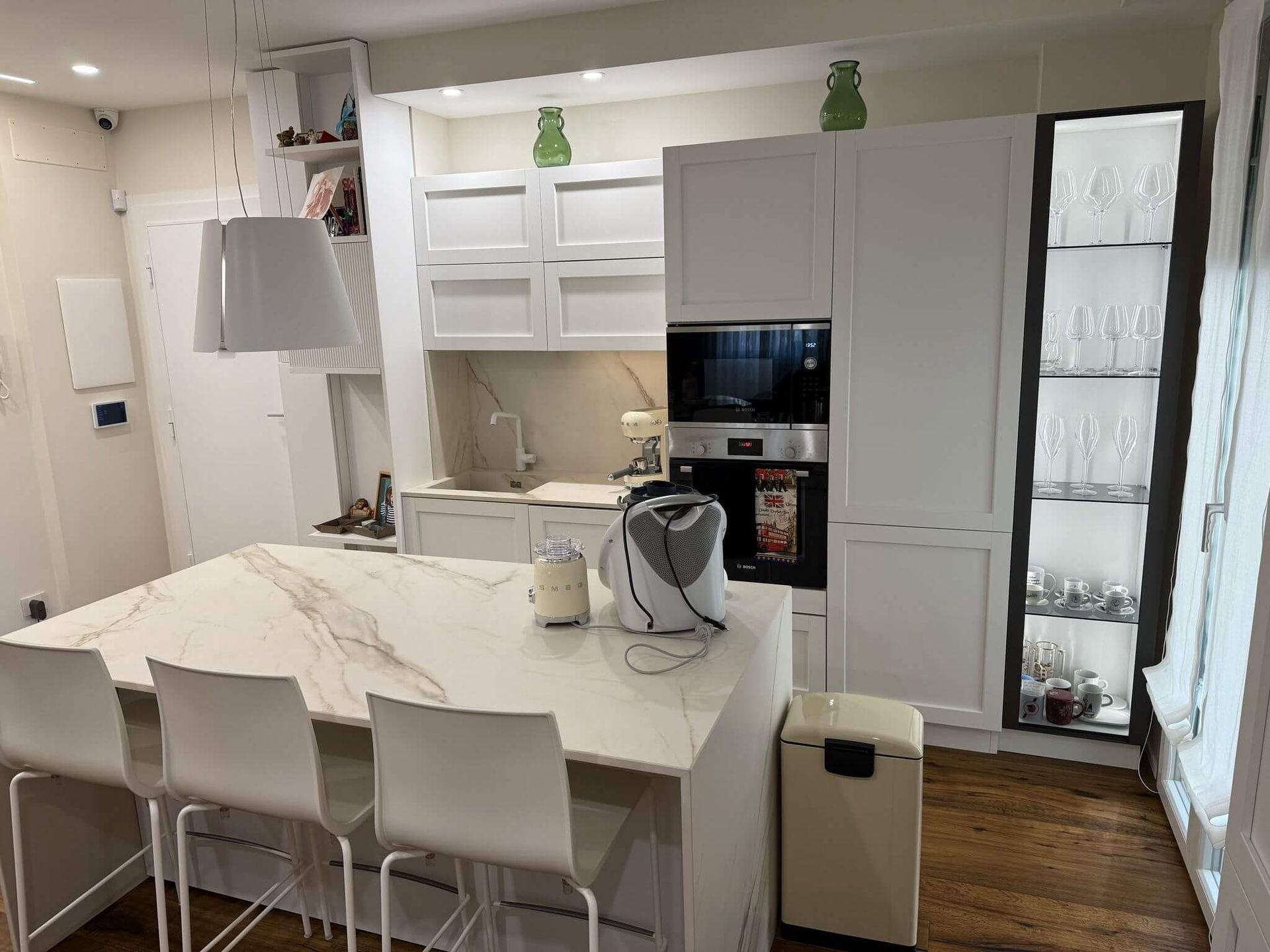 Cucina moderna con isola centrale con piano in marmo bianco, tre sedie bianche e pensili con ante in vetro, su un pavimento in legno scuro.
