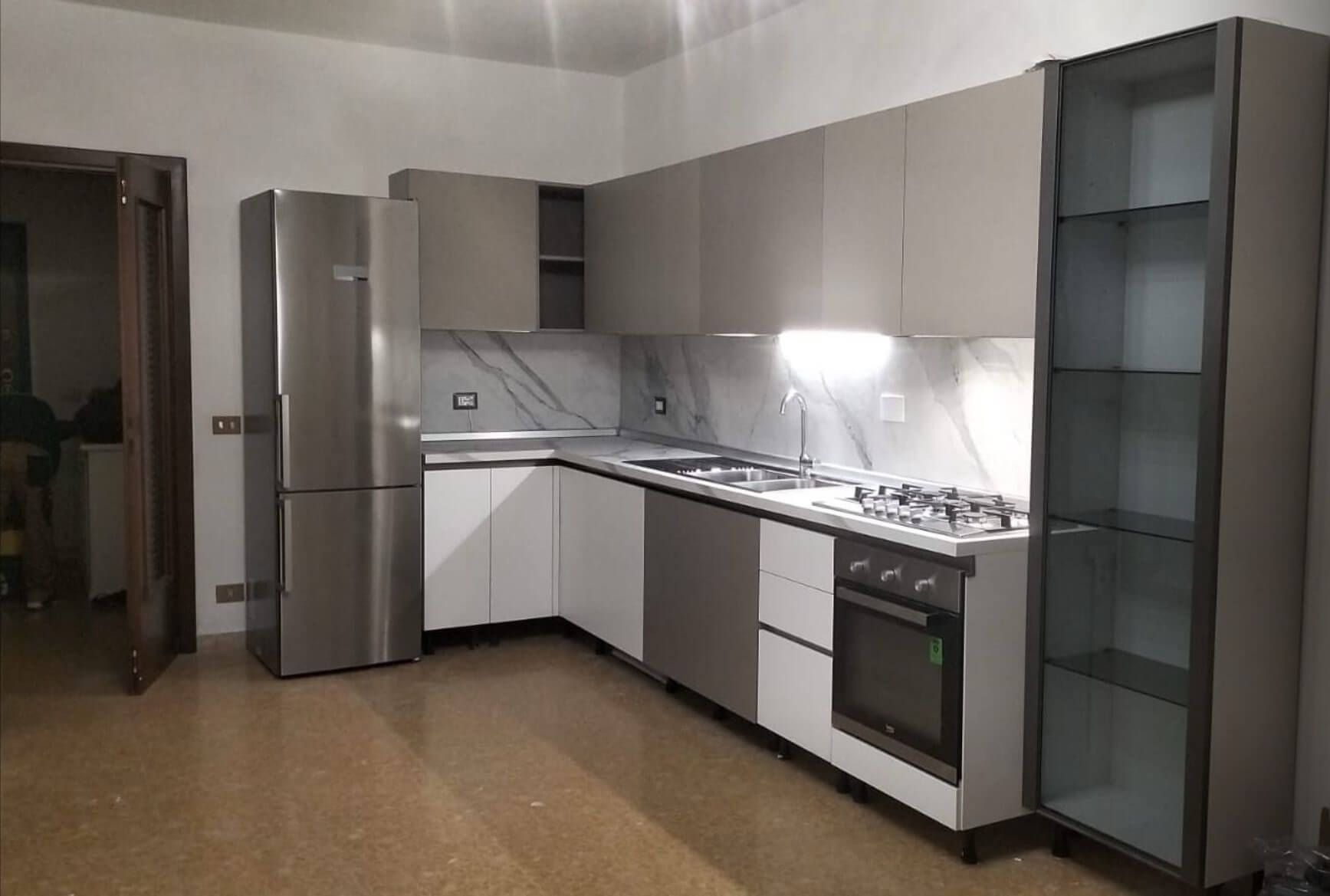 Cucina moderna con frigorifero in acciaio inox, mobili bianchi e tortora, paraschizzi effetto marmo e colonna a vista.
