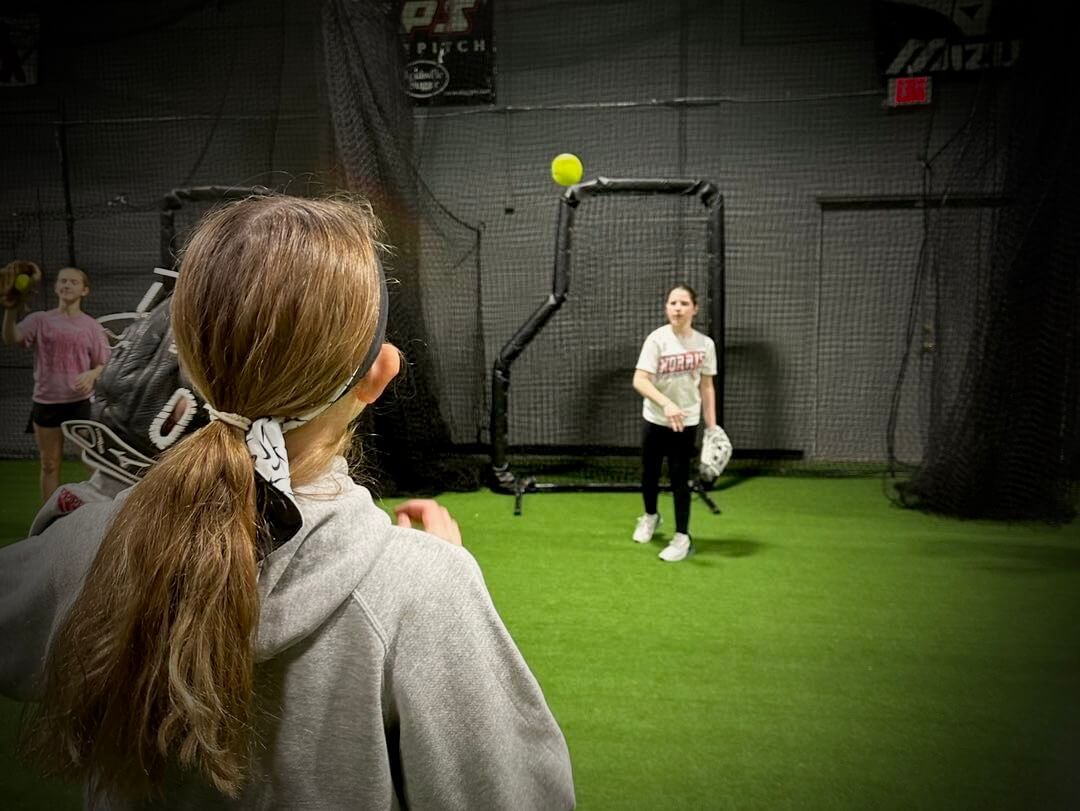 A girl is watching another girl throw a baseball in a cage.
