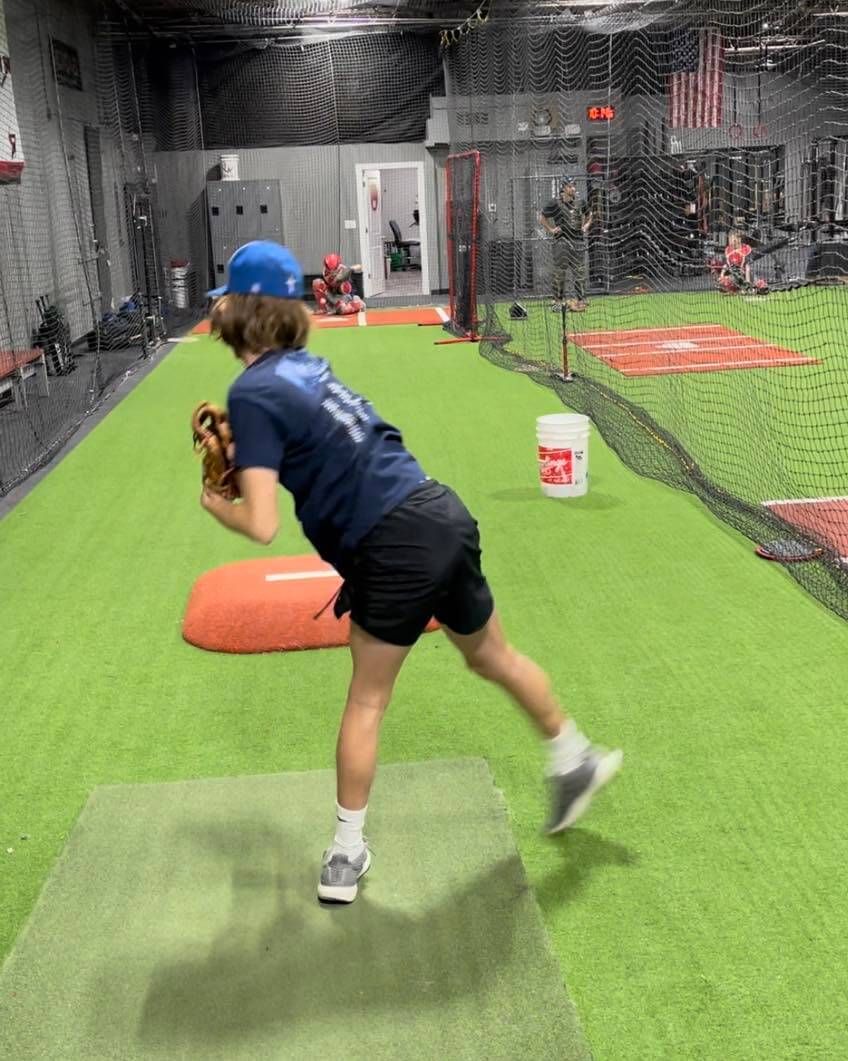 A young baseball pitcher in blue cap and shorts, throwing a ball on a green indoor field.