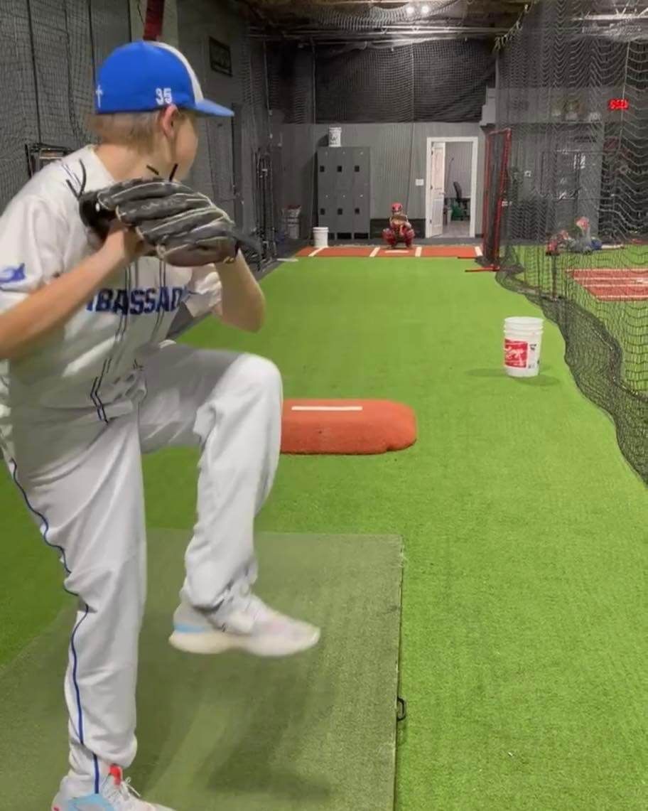 A young baseball pitcher in white uniform with blue cap, poised to throw in an indoor training facility.