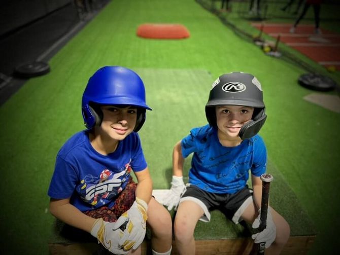 Two young baseball players, wearing helmets and gloves, sit in a batting cage.
