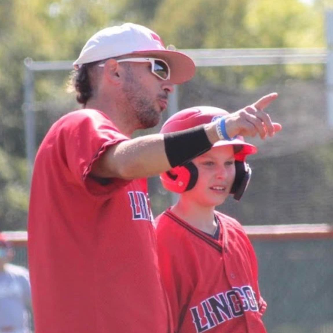 Man in red shirt and cap points, advising a boy in a red baseball helmet.