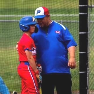 A baseball coach in a blue shirt, cap, and a player in red, helmet, talking on the field.