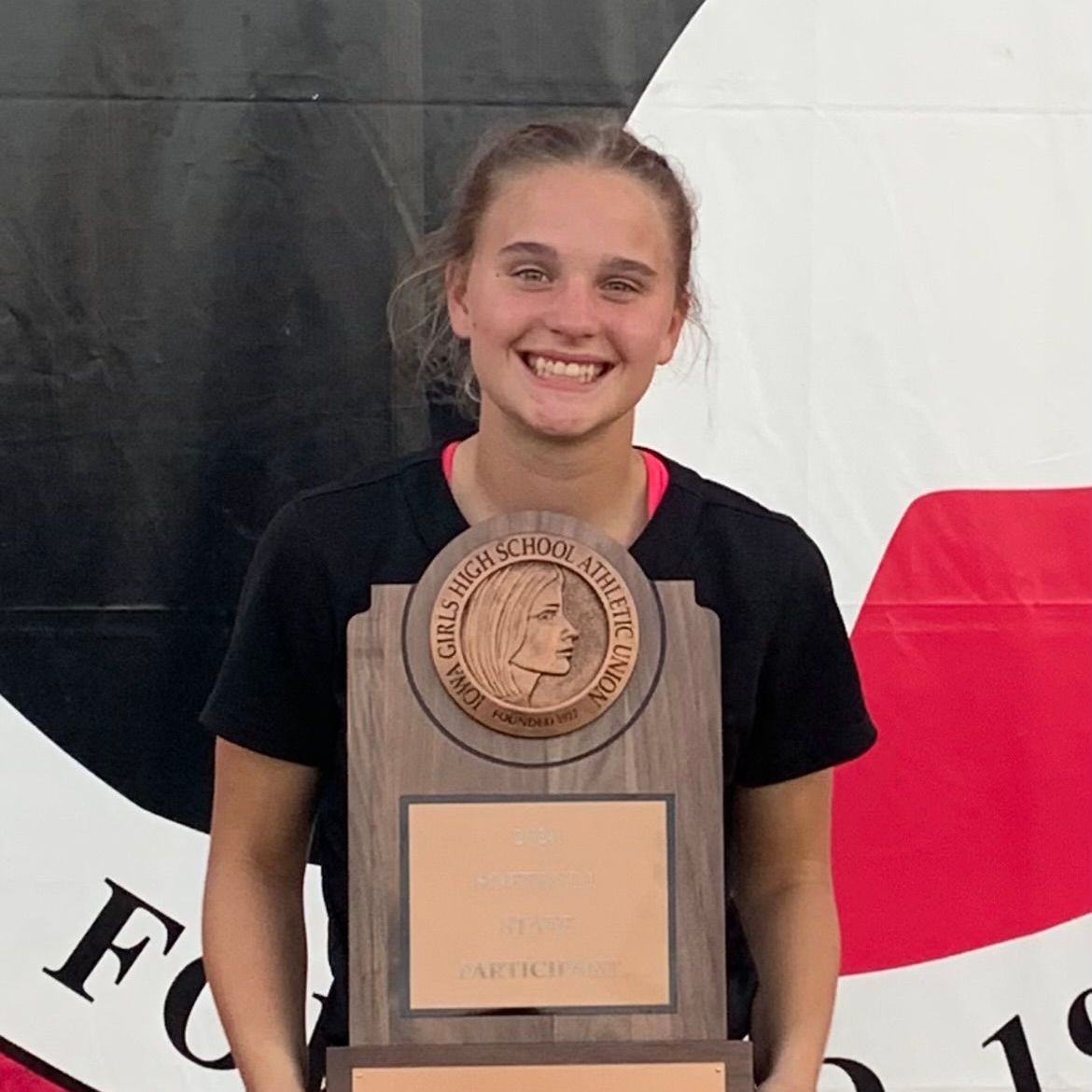 Smiling young woman holding a trophy, standing in front of a black, white, and red backdrop.