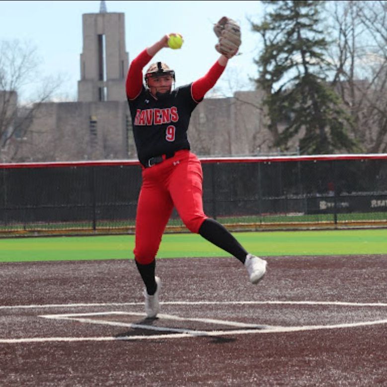 Softball player in red uniform throws ball on a field, with a large building in the background.