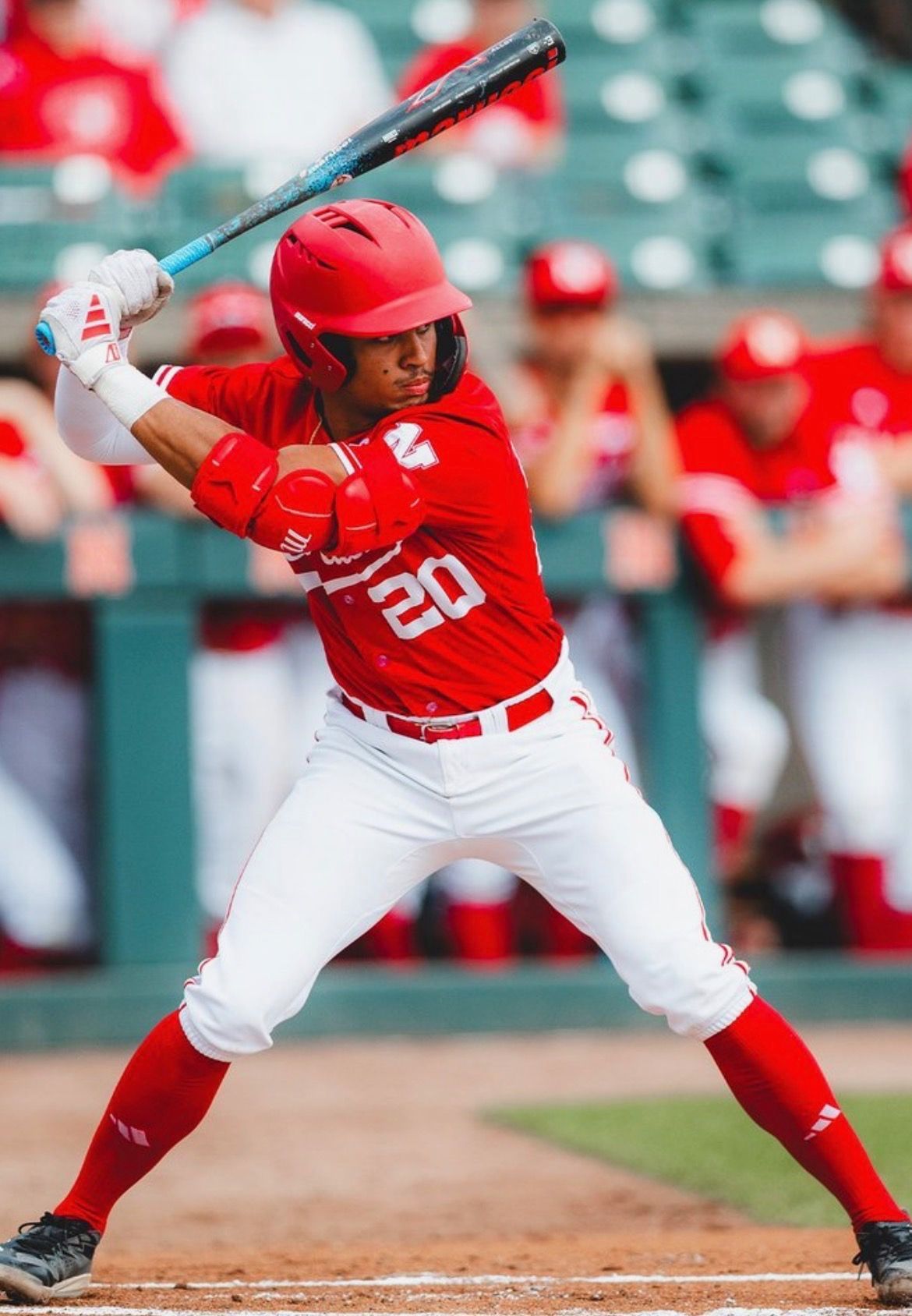 Baseball player in a red uniform, batting stance, holding a bat. White pants, red helmet, field background.