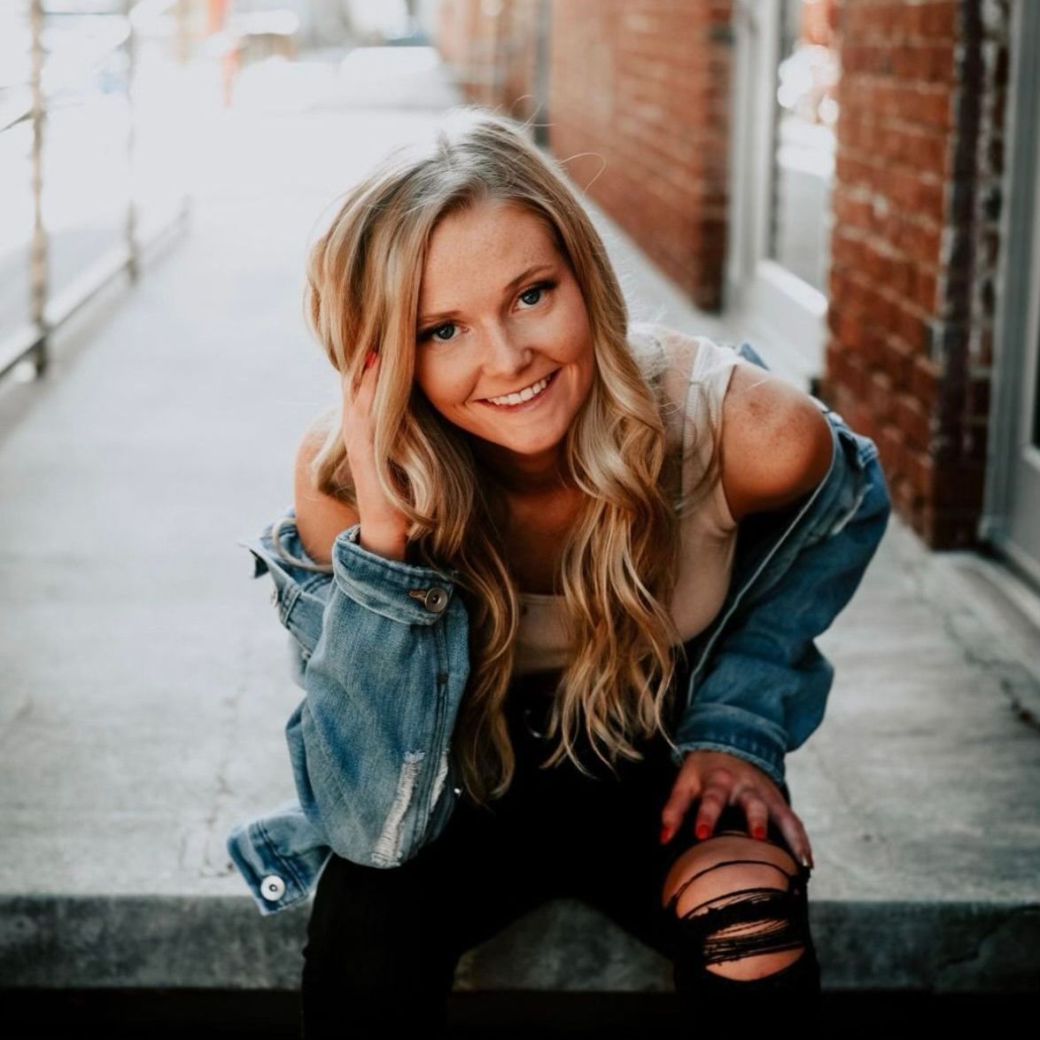 Blonde woman in denim jacket, smiling and leaning against a brick wall.