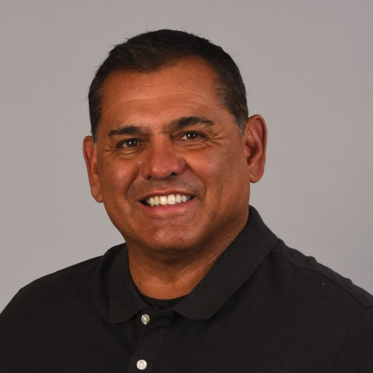 Man in a black polo shirt smiles at the camera, against a gray background.