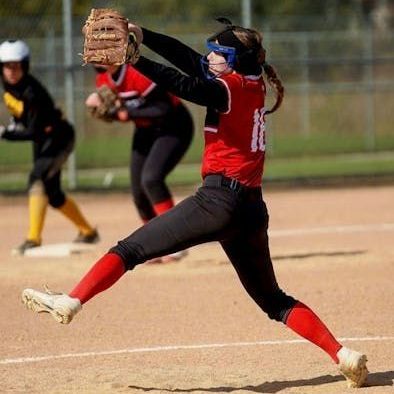 Softball player in red and black uniform pitching, bent at the waist, ball in glove, on a field.
