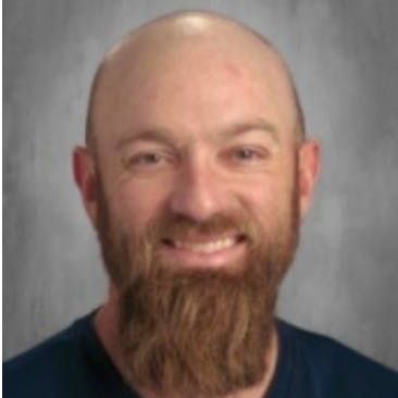 Bald man with beard smiling, wearing a blue shirt against a gray background.