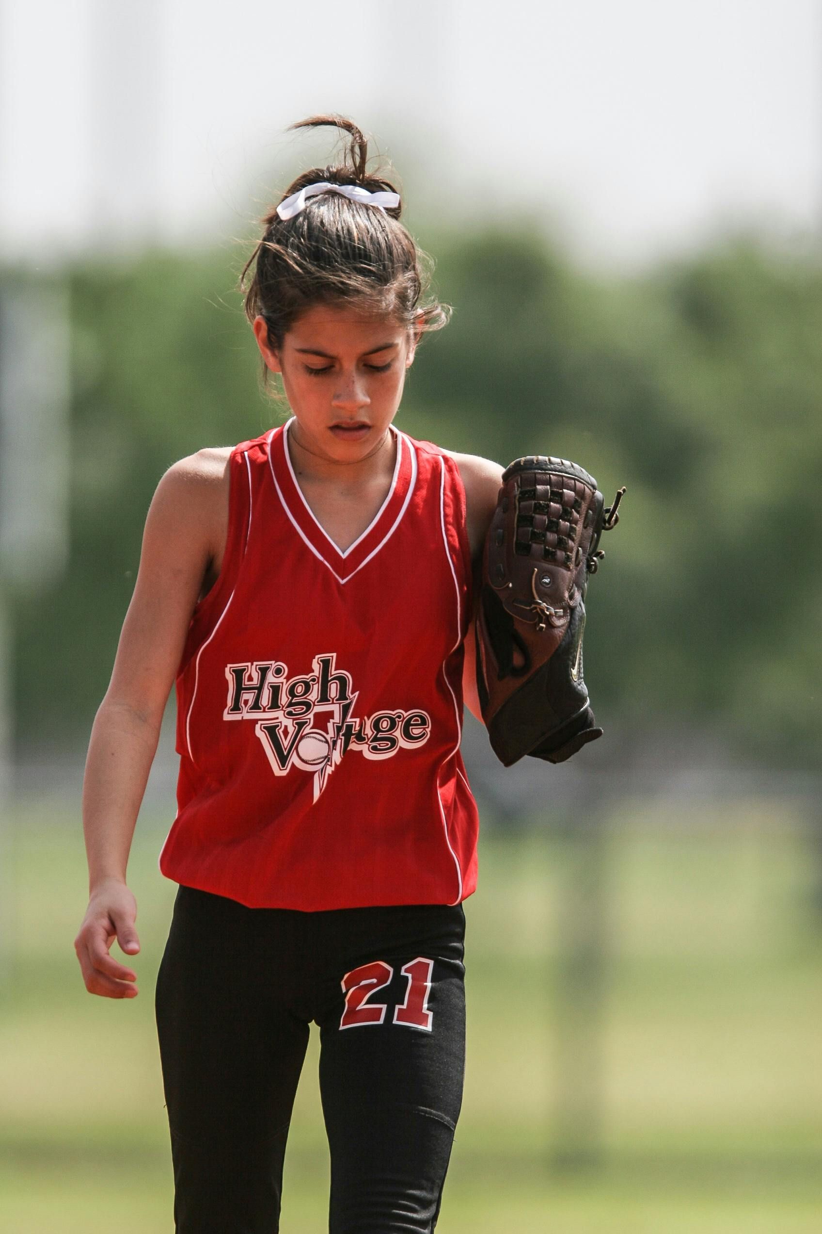 A young girl wearing a red high voltage jersey is holding a baseball glove.