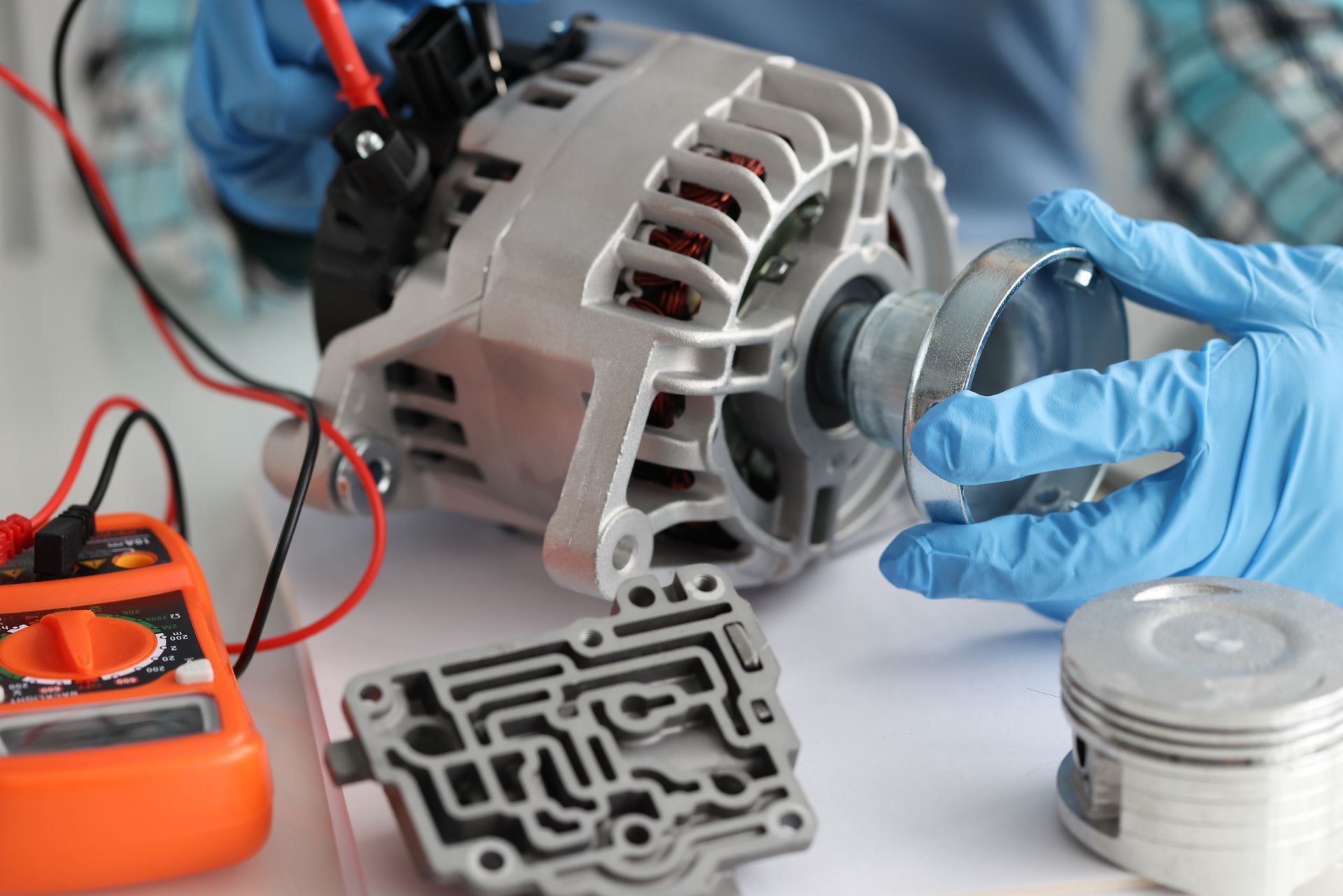 Technician testing an alternator with a multimeter beside disassembled engine parts and gloves | Ralph's Hendersonville Transmission & Auto