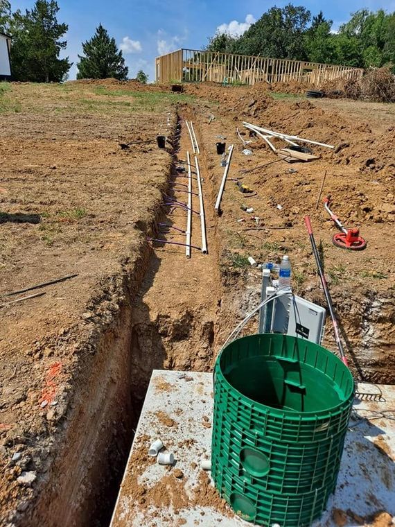 Trenched dirt site with exposed white pipes, green utility box, and scattered construction materials outdoors