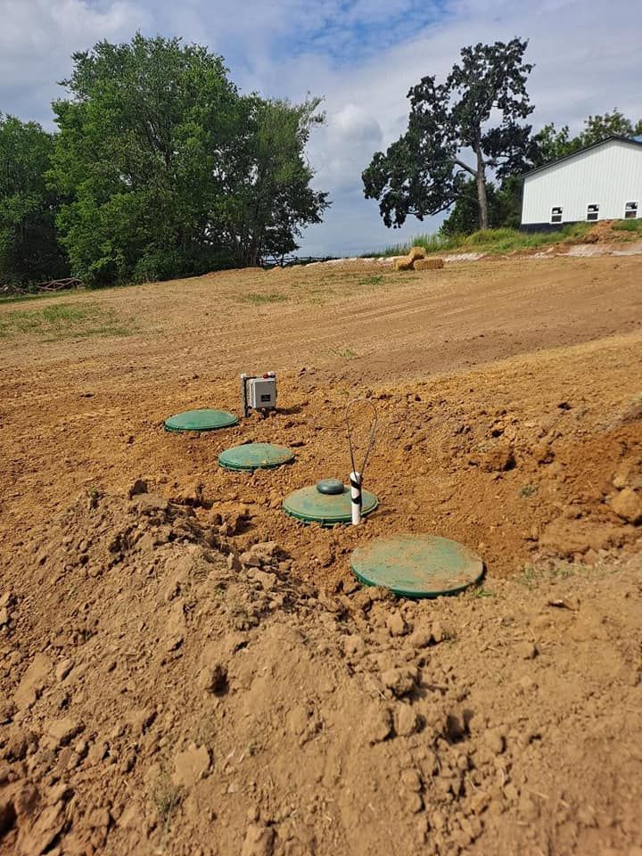 Green utility lids on a dirt hillside with trees and a white building in the background