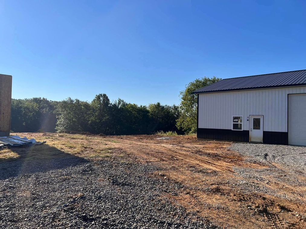 Sunny gravel lot beside a white building with a metal roof and trees in the distance