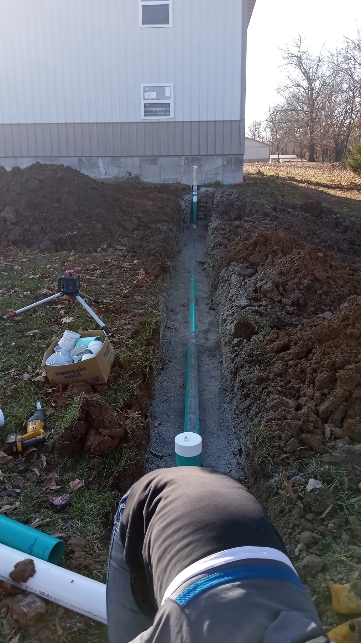 Trench with exposed blue pipe leading to a building foundation, with a white valve in the foreground.