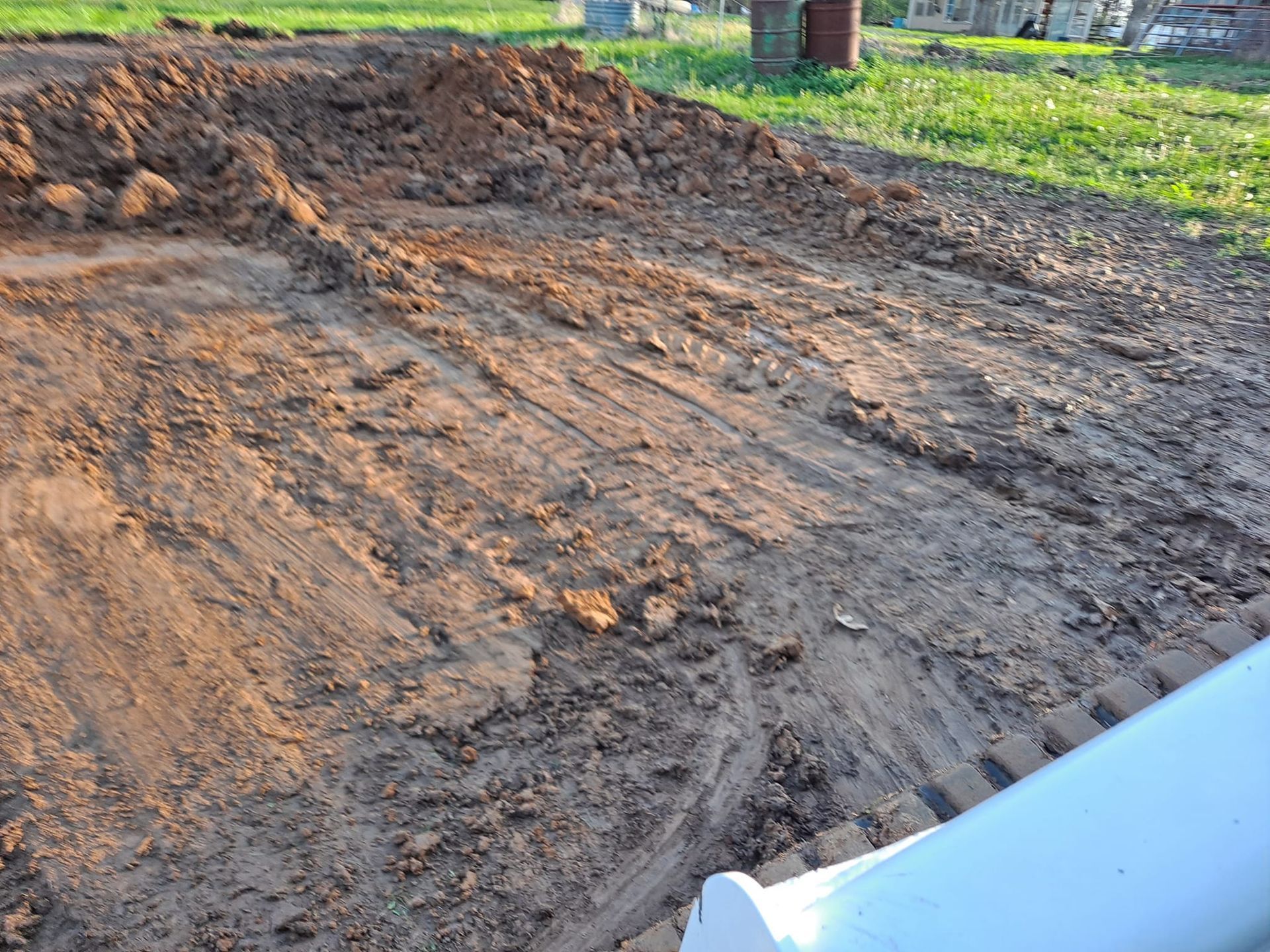 Freshly disturbed muddy ground with tire tracks beside a white vehicle bumper outdoors