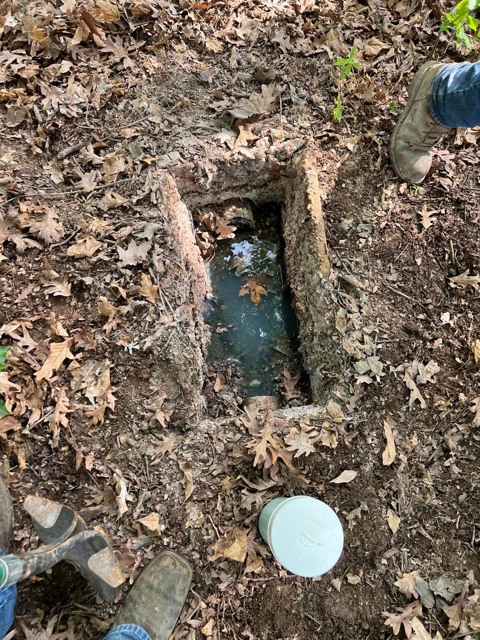 Rectangular hole with standing water in leaf-covered ground, with boots visible around it.