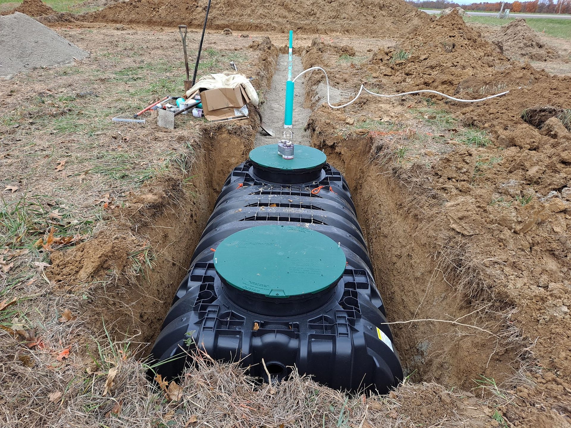 Black septic tank in a dirt trench with green access lids and a white vent pipe during installation
