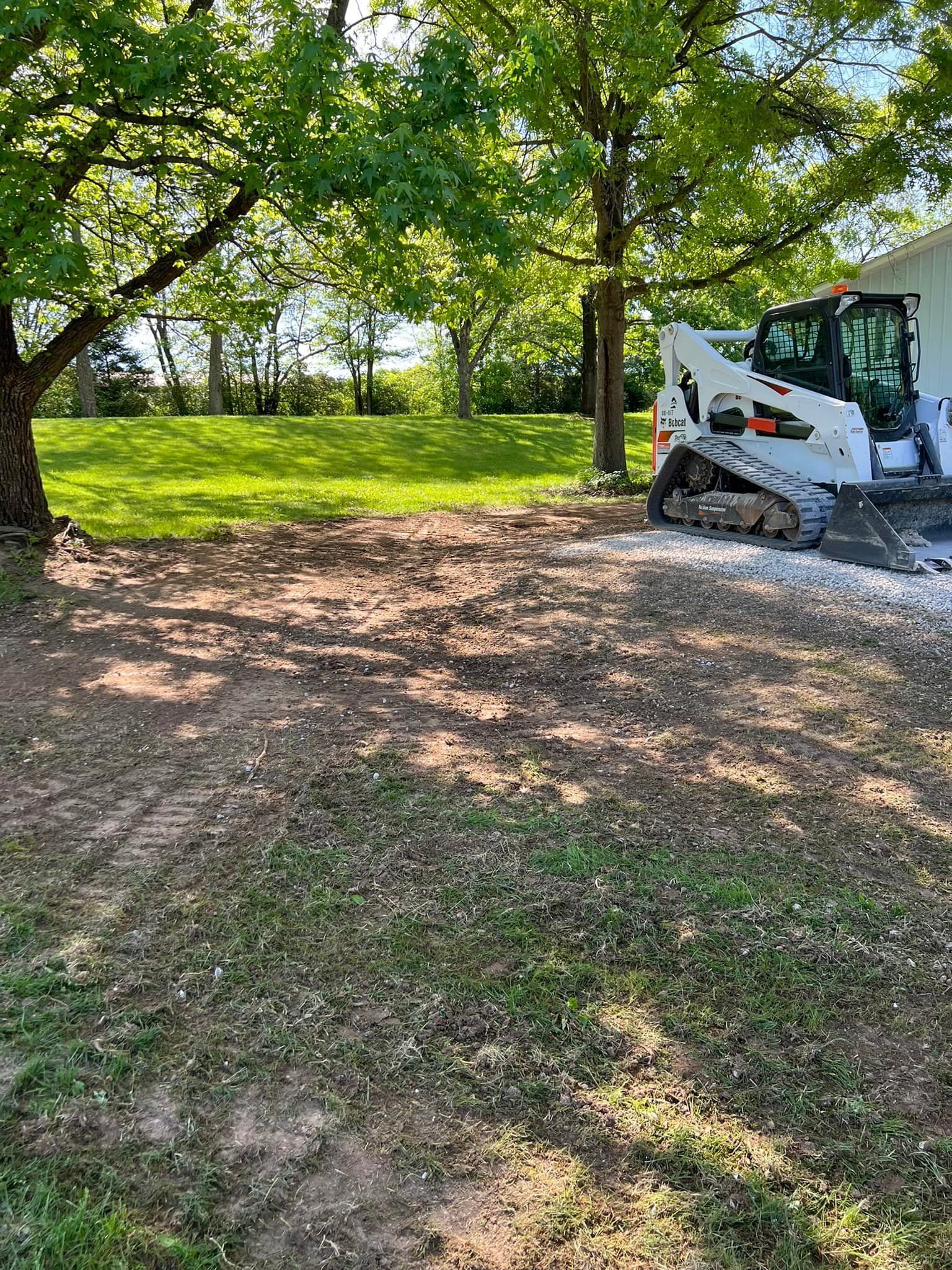 Small white skid steer on a wooded dirt clearing beside a sunlit grassy hill
