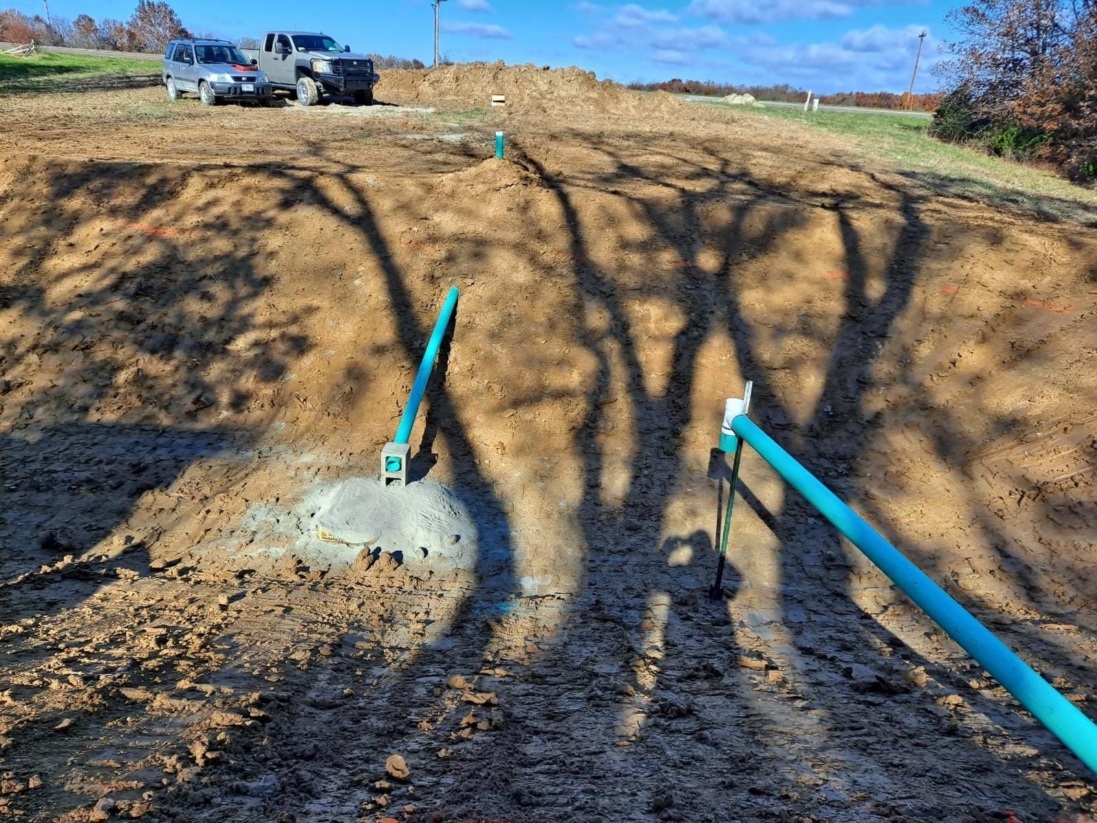 Construction site with blue pipes and utility boxes on a dirt hillside under tree shadows