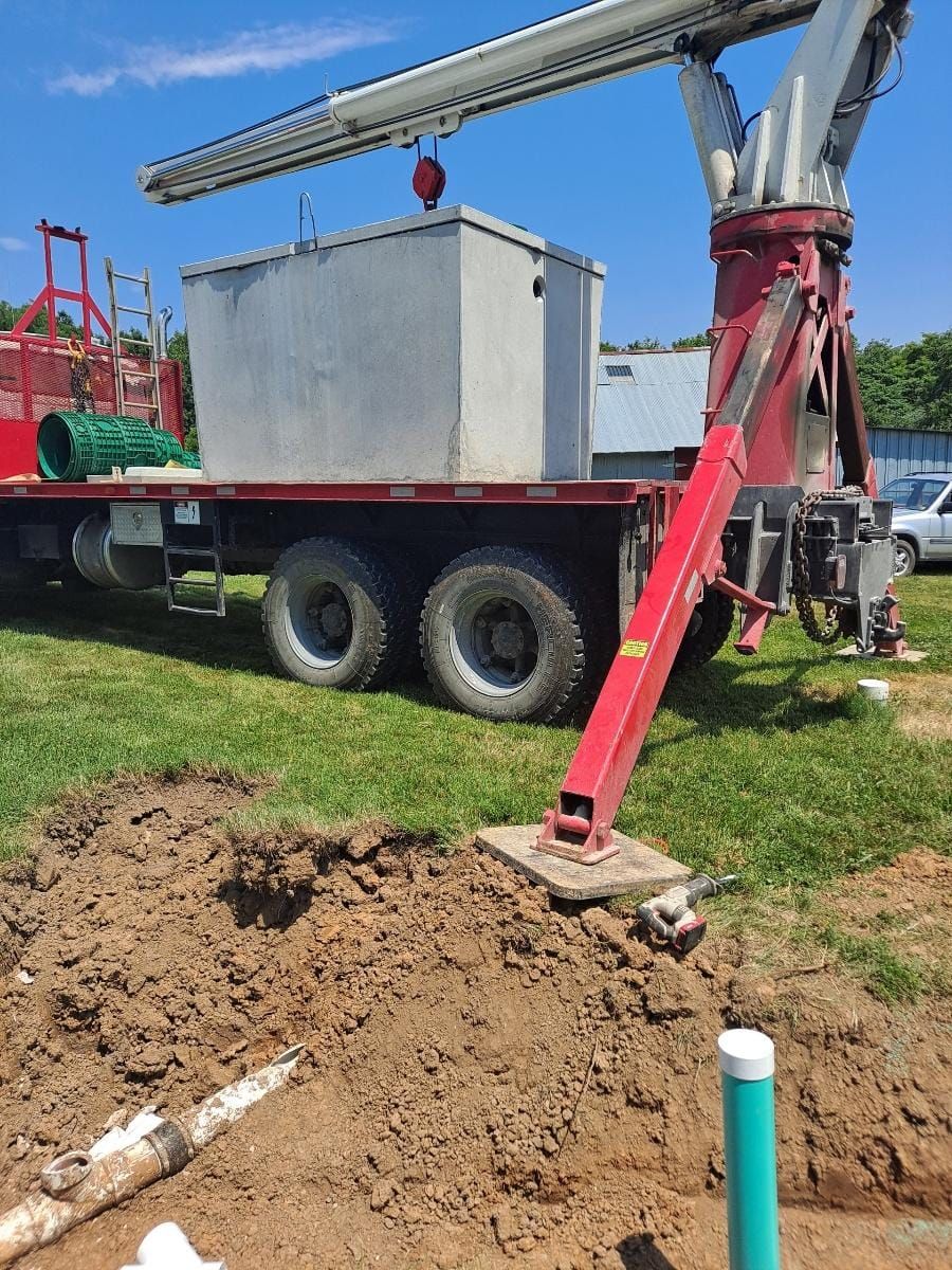Drilling truck beside dirt mound on a grassy site, with red boom and large silver tank under blue sky