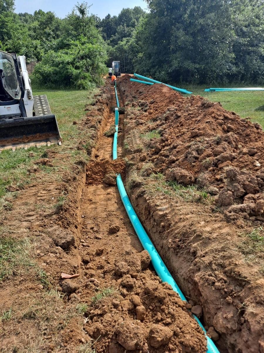Excavator digging a trench with a blue pipe laid along the dirt in a grassy field