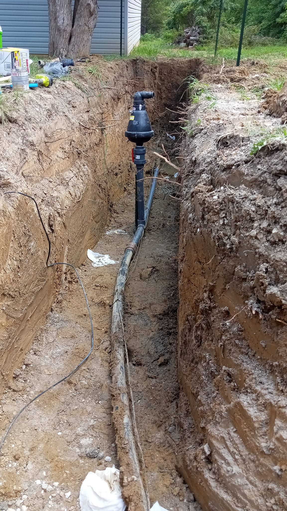Trench with exposed black pipe and valve in muddy soil near a house.