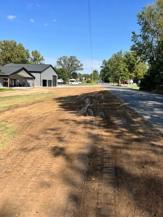 Dirt shoulder beside a paved street in a suburban neighborhood, with houses and trees under a blue sky.