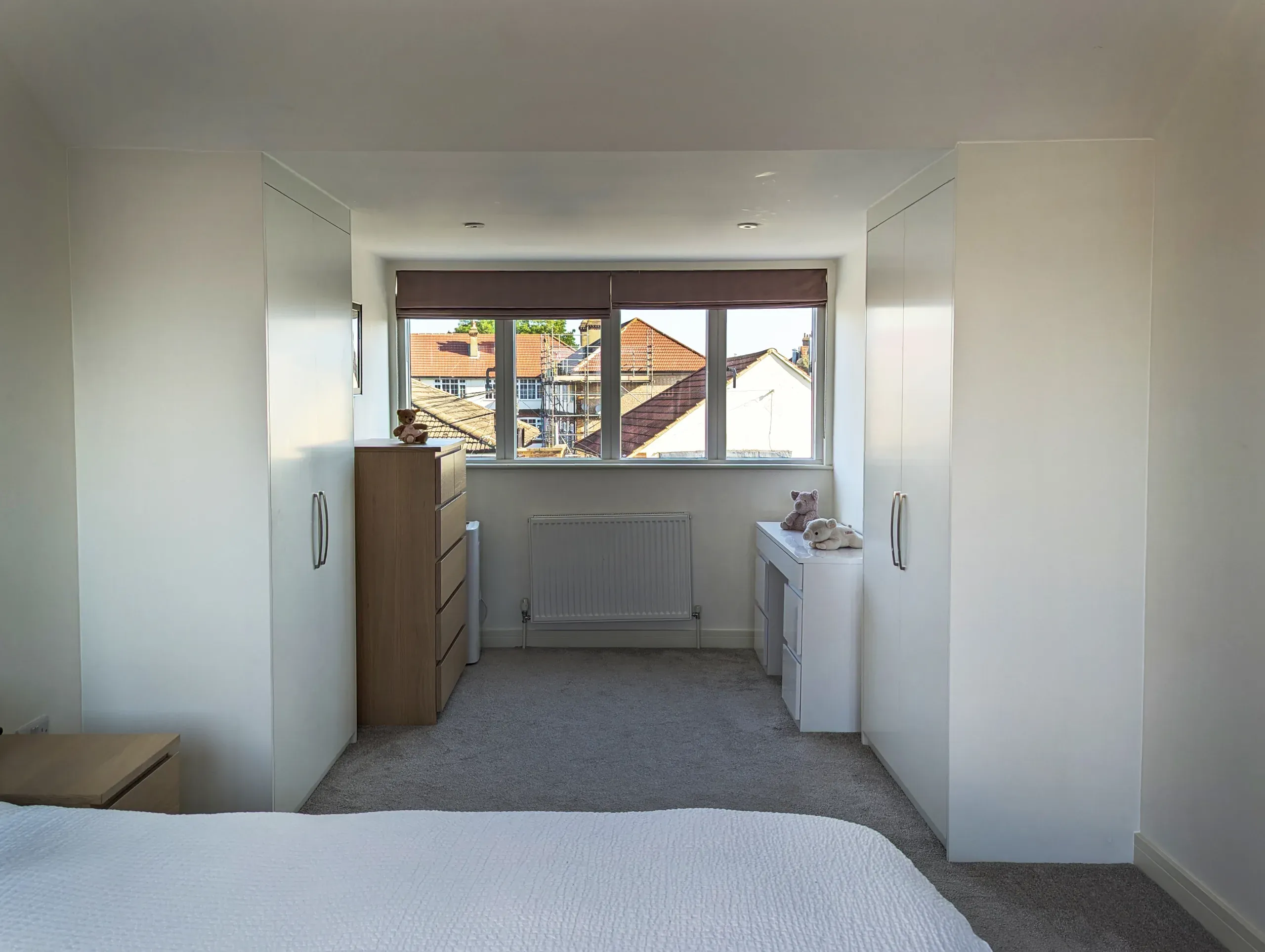 Bedroom with white wardrobes flanking a window, chest of drawers, and a bed.