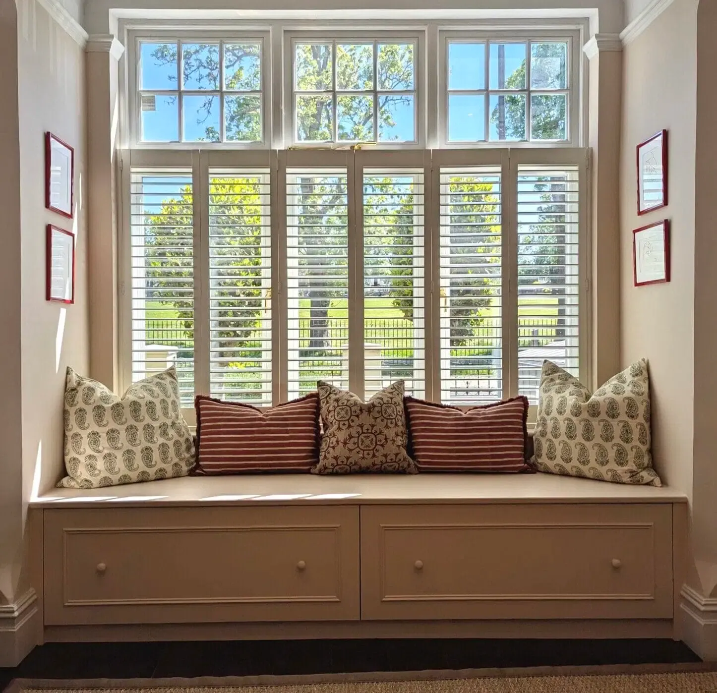 Window seat with shutters, pillows, and framed art. Bright room with a view of a green landscape.