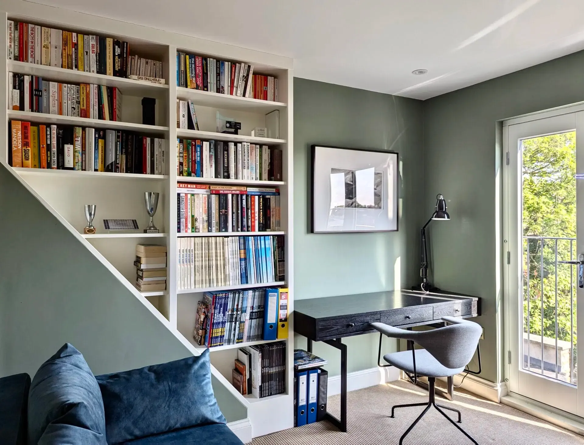 Cozy home office with built-in bookshelves, desk, and French doors to a balcony; green walls, teal couch.