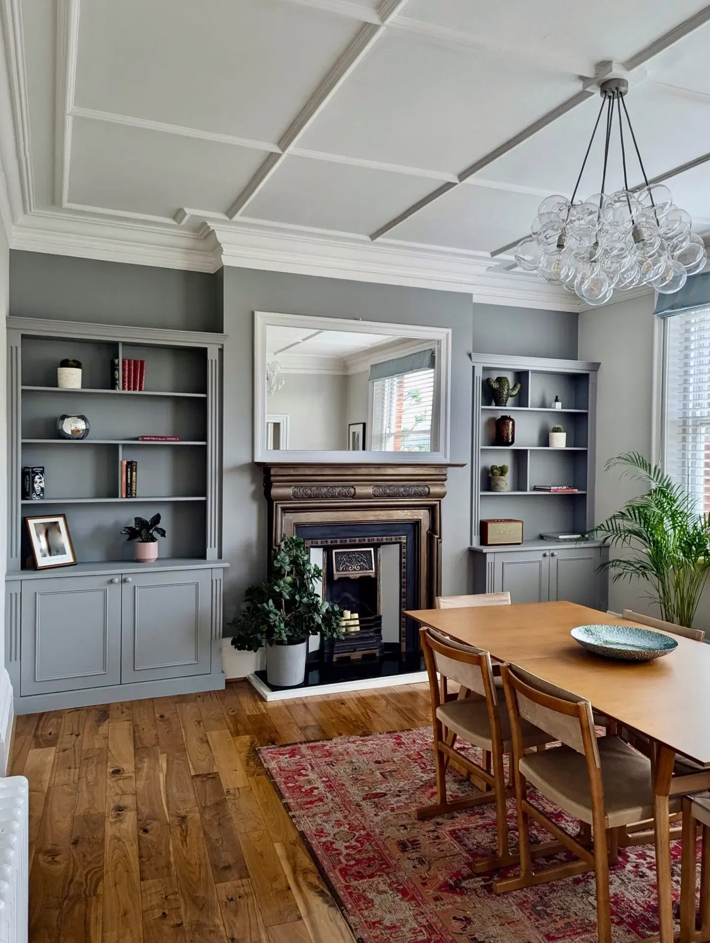 Living room with fireplace, bookshelves, dining table, and rug. Gray walls and wooden floor.