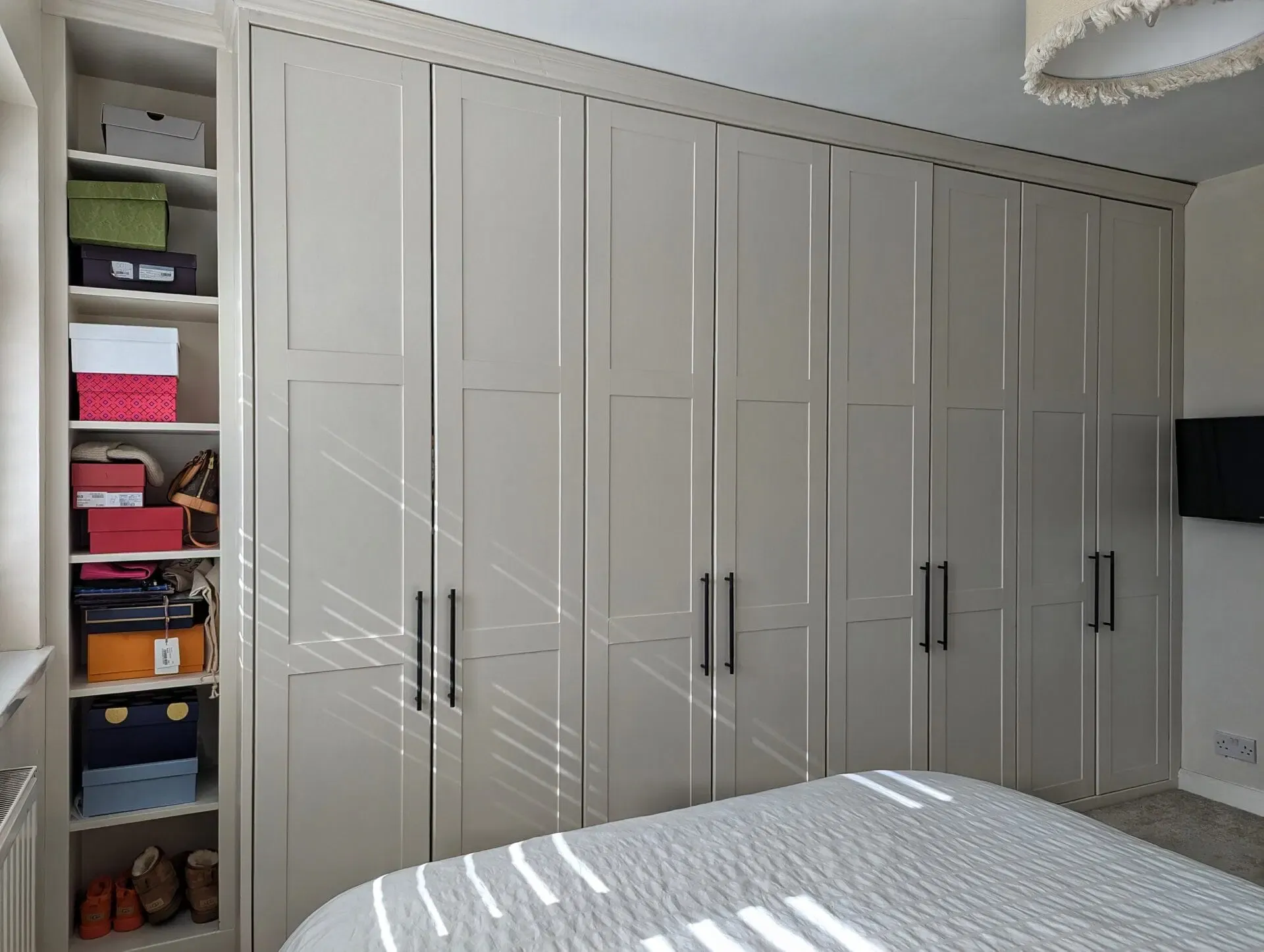 Built-in bedroom wardrobe with light gray doors and a side shelving unit holding shoes and boxes.